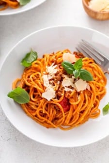 A white plate of spaghetti with tomato meat sauce, topped with parmesan cheese shavings and fresh basil leaves. A fork rests on the side of the plate.