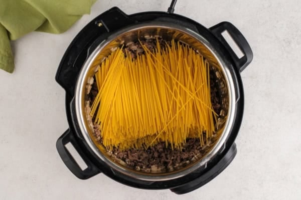 This hearty Instant Pot Spaghetti tastes like it simmered all afternoon, but it's ready in just 30 minutes and made in one single pot. Uncooked spaghetti noodles layered over browned ground beef and onions inside an Instant Pot, viewed from above on a light countertop. A green cloth is partially visible in the top left corner.