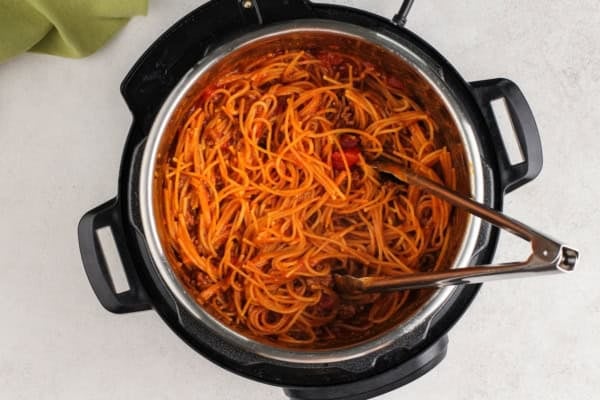 This hearty Instant Pot Spaghetti tastes like it simmered all afternoon, but it's ready in just 30 minutes and made in one single pot. Spaghetti with tomato sauce and ground meat in an electric pressure cooker, with metal tongs resting inside. The cooker is placed on a light-colored countertop.
