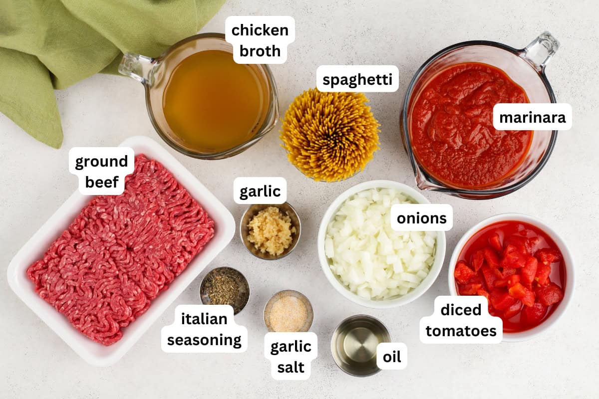 This hearty Instant Pot Spaghetti tastes like it simmered all afternoon, but it's ready in just 30 minutes and made in one single pot. Top-down view of labeled ingredients for spaghetti: ground beef, italian seasoning, garlic salt, garlic, chicken broth, onions, oil, diced tomatoes, marinara sauce, and a bundle of uncooked spaghetti on a white surface.