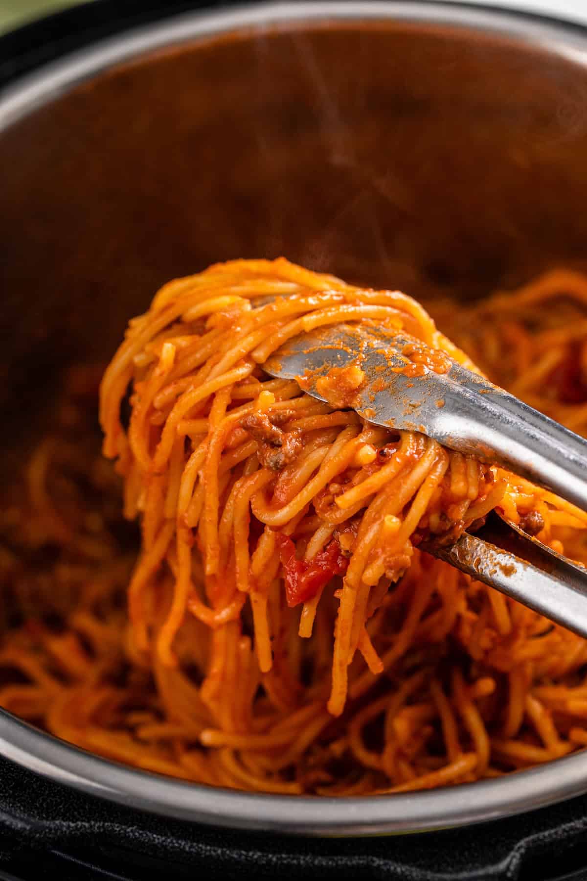 This hearty Instant Pot Spaghetti tastes like it simmered all afternoon, but it's ready in just 30 minutes and made in one single pot. Metal tongs lifting a serving of spaghetti with meat sauce out of a pot, with strands of pasta mixed with ground meat and tomato chunks, and steam rising from the hot dish.
