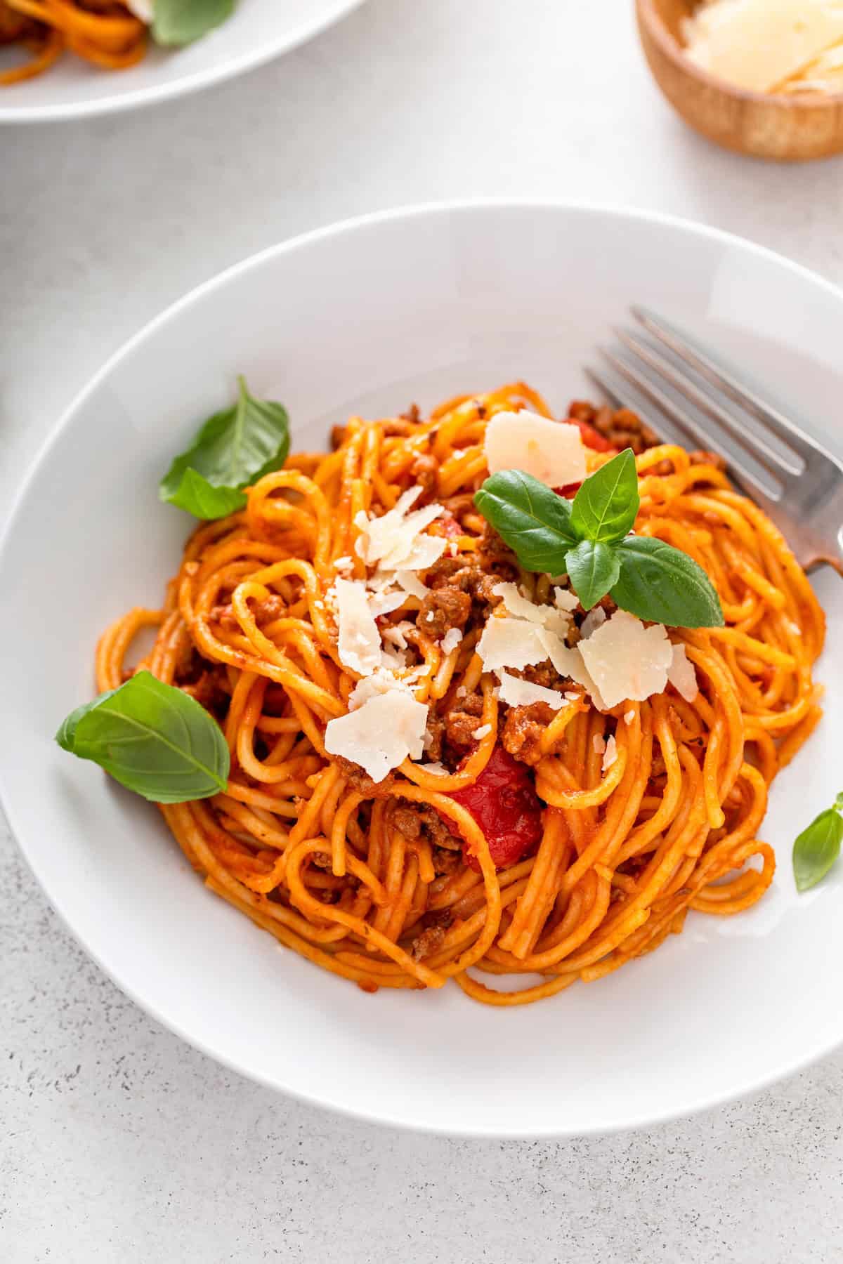 This hearty Instant Pot Spaghetti tastes like it simmered all afternoon, but it's ready in just 30 minutes and made in one single pot. A white plate of spaghetti with tomato meat sauce, topped with parmesan cheese shavings and fresh basil leaves. A fork rests on the side of the plate.