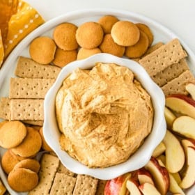 A white plate holds a bowl of marshmallow sweet potato pie dip, surrounded by apple slices, graham crackers, and vanilla wafer cookies on a yellow polka dot cloth.