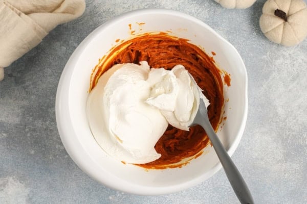 This no-bake Pumpkin Dip is perfectly spiced and made with marshmallow fluff to keep it extra fluffy. Party ready in just 5 minutes! A white bowl with seasoned pumpkin puree and a large dollop of marshmallow fluff and cool whip, being mixed with a gray spatula.
