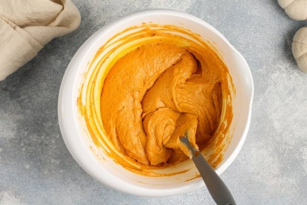 This no-bake Pumpkin Dip is perfectly spiced and made with marshmallow fluff to keep it extra fluffy. Party ready in just 5 minutes! A white bowl filled with creamy, orange pumpkin pie dip is shown on a light countertop. A spatula rests in the bowl.