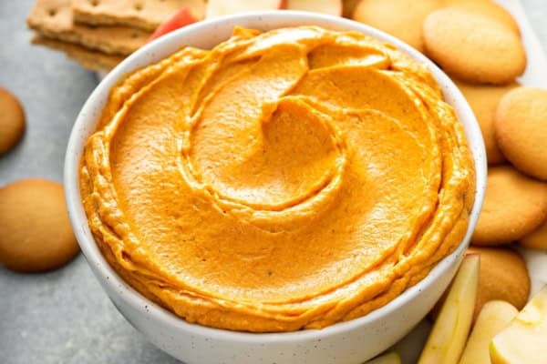 This no-bake Pumpkin Dip is perfectly spiced and made with marshmallow fluff to keep it extra fluffy. Party ready in just 5 minutes! A white bowl filled with creamy orange pumpkin pie dip sits on a tray, surrounded by Nilla wafer cookies, graham crackers, and apple slices.