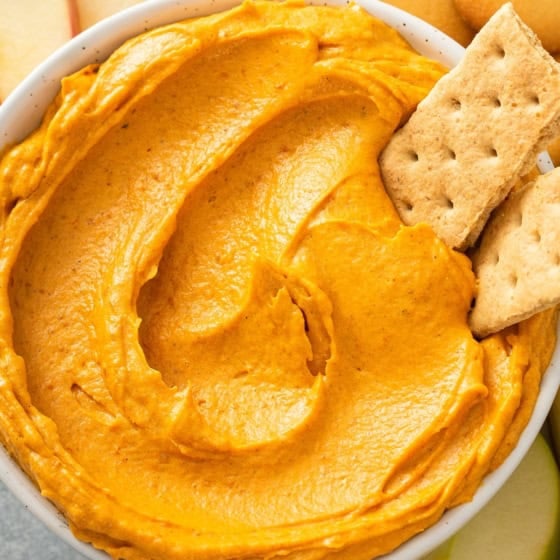 A bowl of creamy orange pumpkin dip is surrounded by apple slices, Nilla wafer cookies, and two graham crackers dipped into the dip.