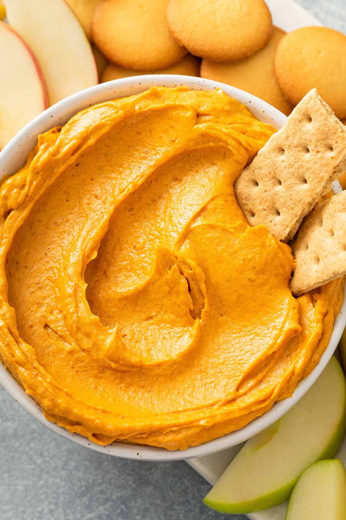This no-bake Pumpkin Dip is perfectly spiced and made with marshmallow fluff to keep it extra fluffy. Party ready in just 5 minutes! A bowl of creamy orange pumpkin dip is surrounded by apple slices, Nilla wafer cookies, and two graham crackers dipped into the dip.