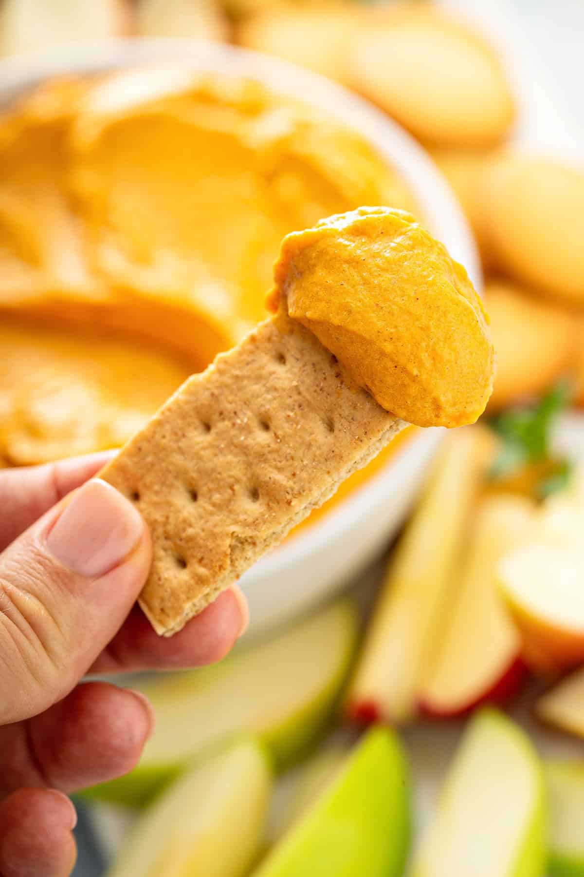 This no-bake Pumpkin Dip is perfectly spiced and made with marshmallow fluff to keep it extra fluffy. Party ready in just 5 minutes! A hand holds a graham cracker topped with orange pumpkin dip. A bowl of pumpkin pie dip and sliced apples are blurred in the background.