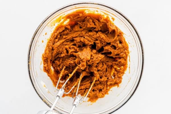This Sweet Potato Pie Dip tastes like a classic sweet potato pie, but in a scoopable form. Serve it with gingersnaps or graham crackers! A glass bowl filled with mashed sweet potato and spices being mixed with an electric hand mixer on a white surface.