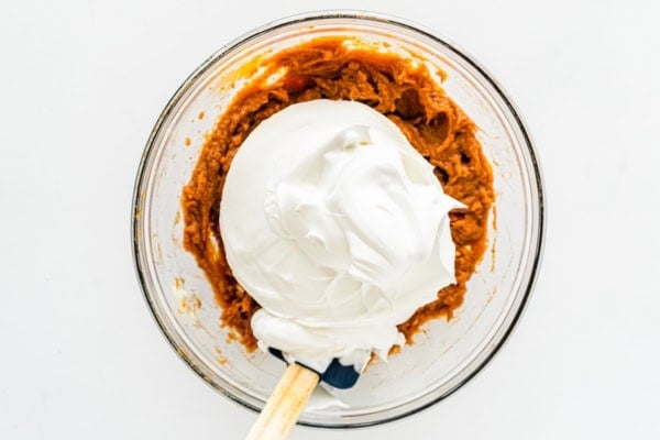 This Sweet Potato Pie Dip tastes like a classic sweet potato pie, but in a scoopable form. Serve it with gingersnaps or graham crackers! A glass bowl with mashed sweet potatoes and a large dollop of marshmallow fluff and cool whip on top. A spatula rests inside the bowl, ready to mix the ingredients together.