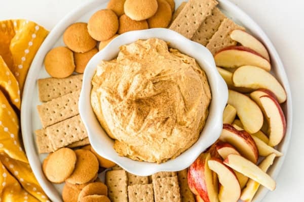 This Sweet Potato Pie Dip tastes like a classic sweet potato pie, but in a scoopable form. Serve it with gingersnaps or graham crackers! A bowl of creamy sweet potato pie dip is surrounded by graham crackers, vanilla wafers, and apple slices on a white plate, with a yellow napkin to the side.