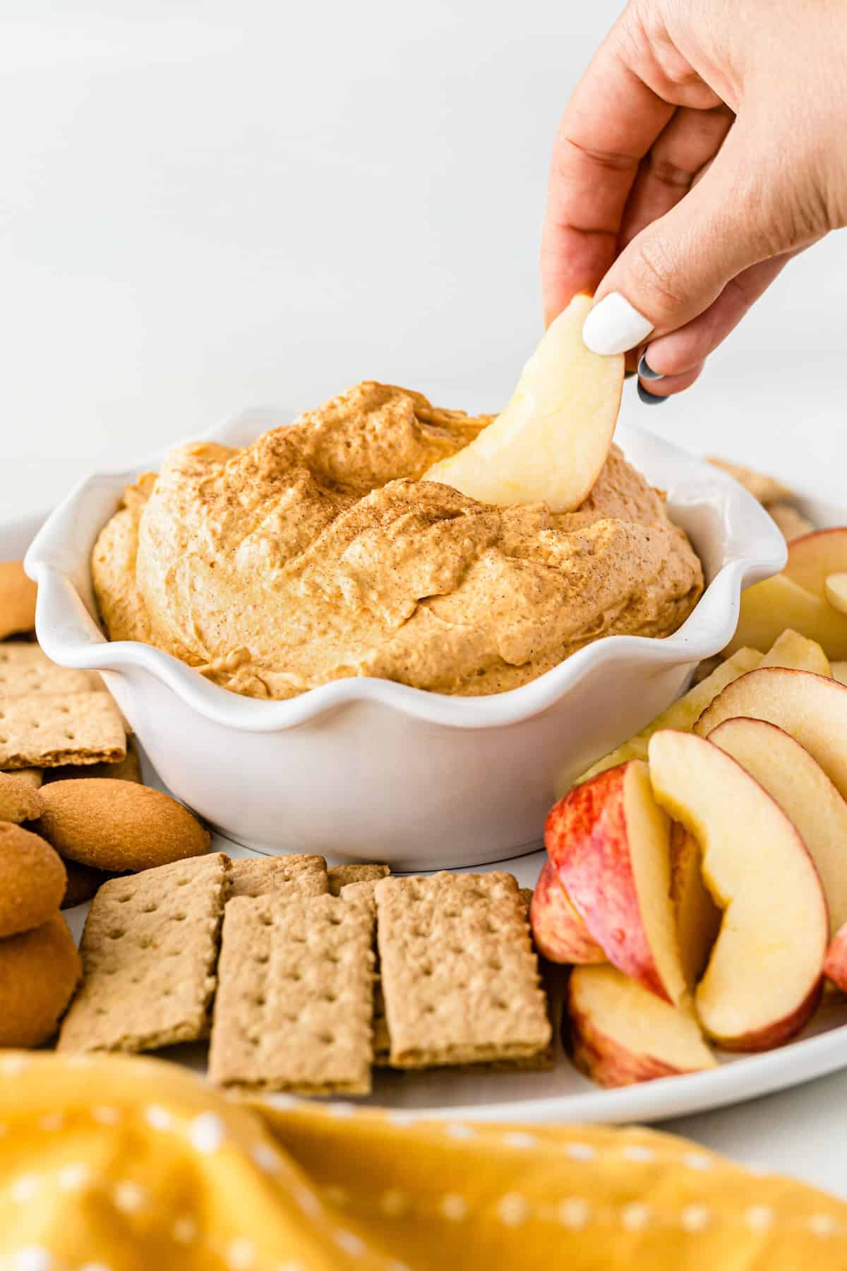 This Sweet Potato Pie Dip tastes like a classic sweet potato pie, but in a scoopable form. Serve it with gingersnaps or graham crackers! A hand dips an apple slice into a bowl of creamy sweet potato pie dip, surrounded by graham crackers, vanilla wafers, and more apple slices on a white tray.