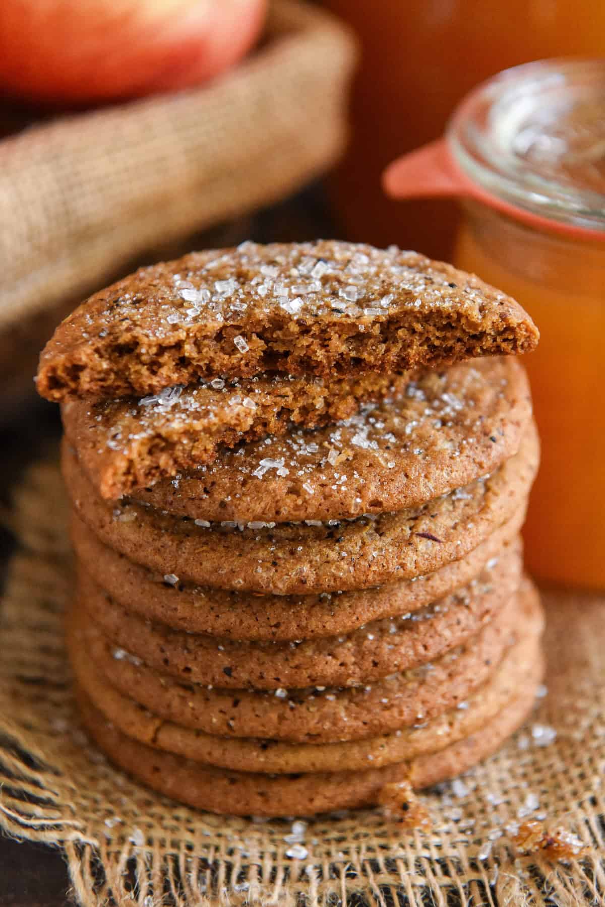 So soft and chewy, these Apple Cider Cookies are made with instant apple cider mix for big cider flavor with no stovetop reduction needed! A stack of chewy apple cider cookies topped with coarse sugar sits on burlap. The top cookie is broken in half, showing its soft center. A jar of apple cider and an apple are blurred in the background.
