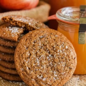 A stack of apple cider cookies with sugar crystals sits on burlap. One cookie rests in front. Behind them are a red apple and a jar of apple cider.