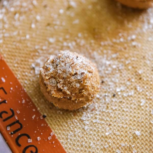 So soft and chewy, these Apple Cider Cookies are made with instant apple cider mix for big cider flavor with no stovetop reduction needed! A round cookie dough ball topped with a coarse sugar and apple cider instant drink mixture sits on a baking mat. Part of another dough ball and an orange border with partial text are visible in the background.