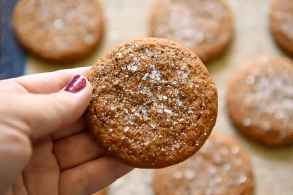 So soft and chewy, these Apple Cider Cookies are made with instant apple cider mix for big cider flavor with no stovetop reduction needed! A hand holds an apple cider cookie topped with coarse sugar. More cookies are visible on a baking tray in the background.