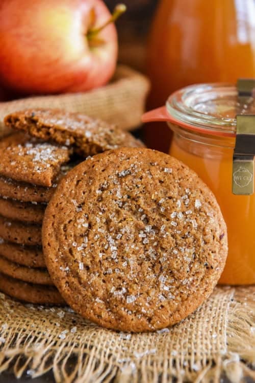 A stack of apple cider cookies with sugar crystals sits on burlap. One cookie rests in front. Behind them are a red apple and a jar of apple cider.