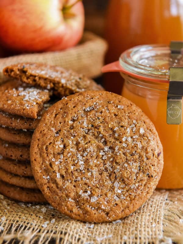 A stack of apple cider cookies with sugar crystals sits on burlap. One cookie rests in front. Behind them are a red apple and a jar of apple cider.