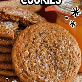 A close-up of a stack of apple cider cookies topped with sugar crystals, with a glass of cider and an apple in the background.