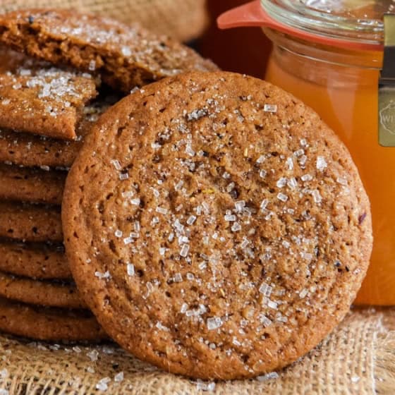 A stack of sugar-sprinkled apple cider cookies sits on burlap next to a jar of golden liquid, likely apple cider, with cinnamon sticks nearby.
