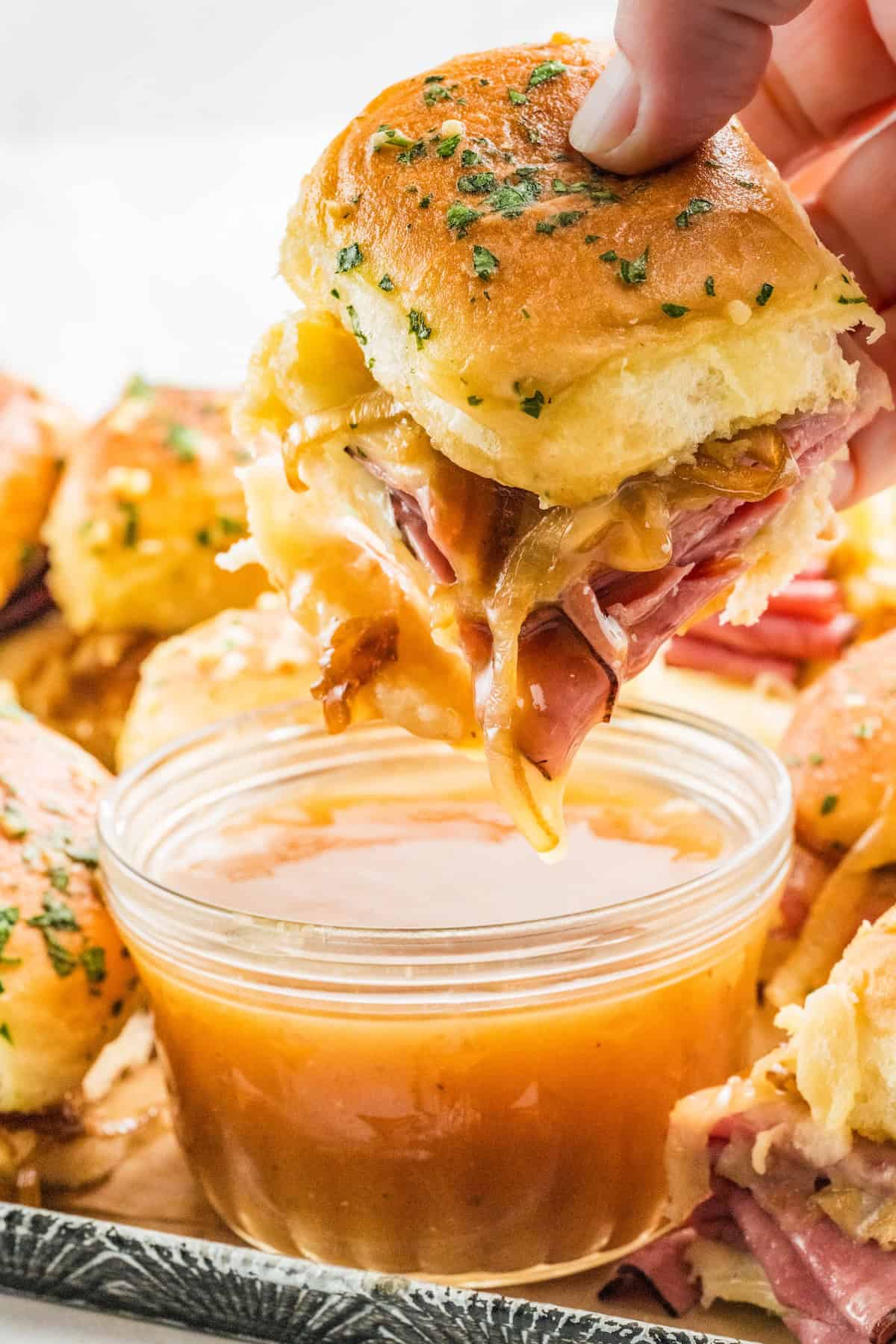 A hand dips a cheesy French dip slider on a golden bun with herbs into a bowl of au jus sauce. Other roast beef sliders are visible in the background.
