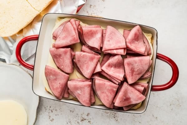 Rectangular baking dish with stacked slices of folded roast beef deli meat and cheese on top of bread, ready to be baked.