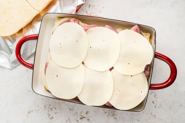 A rectangular baking dish with layered roast beef deli meat and six slices of provolone cheese on top of slider buns, ready to be baked.