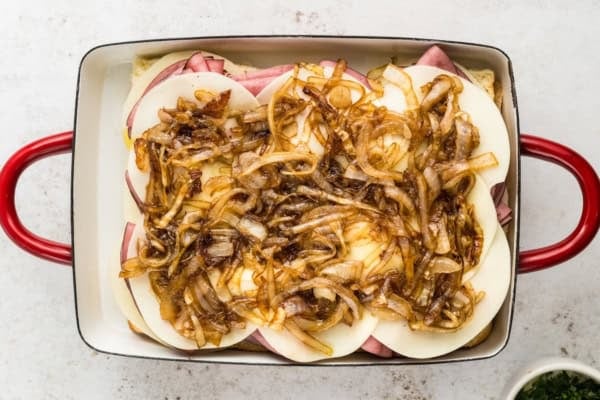 A casserole dish with layers of slider rolls, roast beef, provolone cheese, and caramelized onions, ready to be baked.