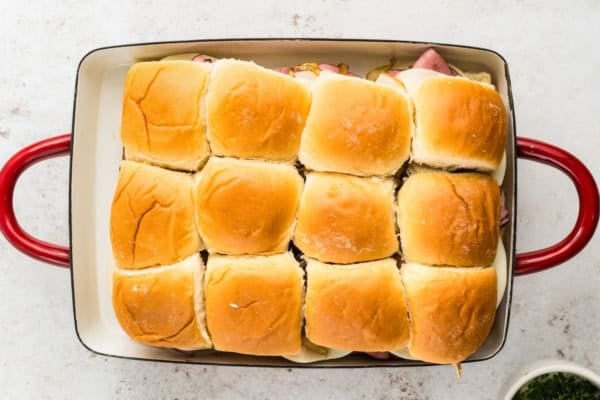A baking dish with red handles contains a batch of twelve slider sandwiches arranged in three rows of four.