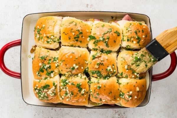 A baking dish with 12 slider buns topped with fresh herb and garlic butter, being brushed with more butter using a pastry brush.