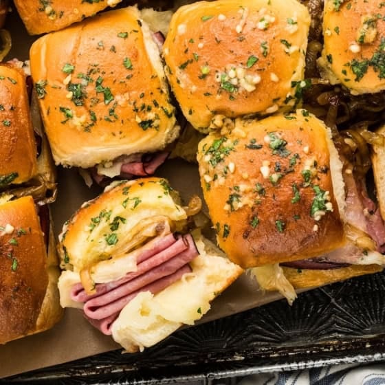 A tray of French dip sliders on golden, herb-topped buns, with melted cheese and slices of roast beef and cheese visible inside.
