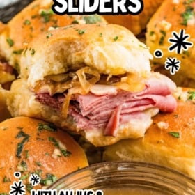 A close-up of French dip sliders topped with herbs, filled with layers of roast beef and caramelized onions, with a dish of au jus in the foreground.