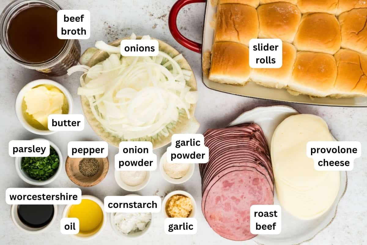 Top-down view of ingredients for French dip sliders recipe: slider rolls, sliced roast beef, provolone cheese, sliced onions, butter, oil, parsley, pepper, onion powder, garlic powder, garlic, cornstarch, Worcestershire sauce, beef broth.
