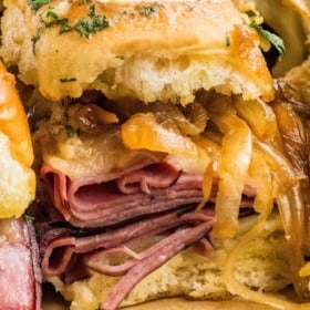 Close-up of a French dip slider with layers of roast beef, caramelized onions, melted cheese, and herbs on a toasted bun.