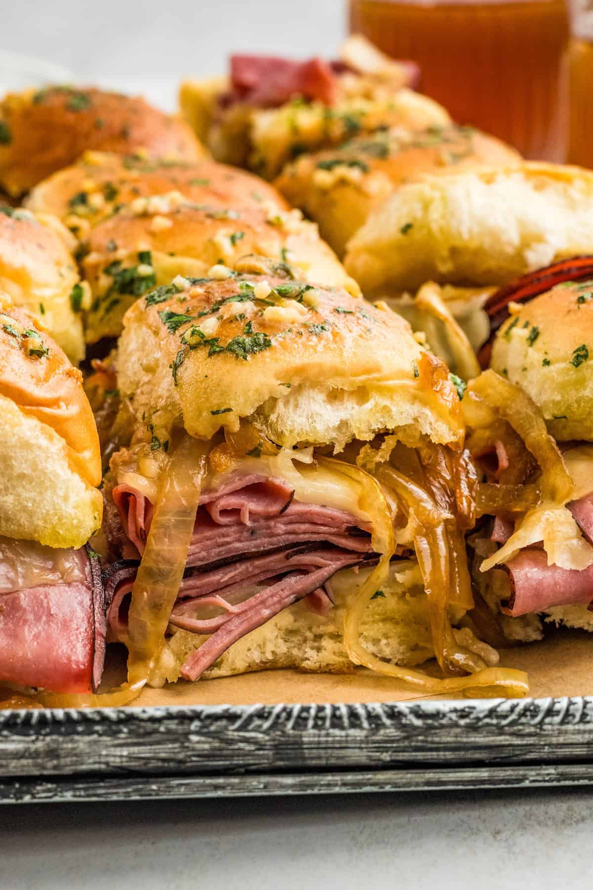 A close-up of French dip roast beef sliders filled with melted provolone cheese, and caramelized onions on a tray, topped with herbed garlic butter.
