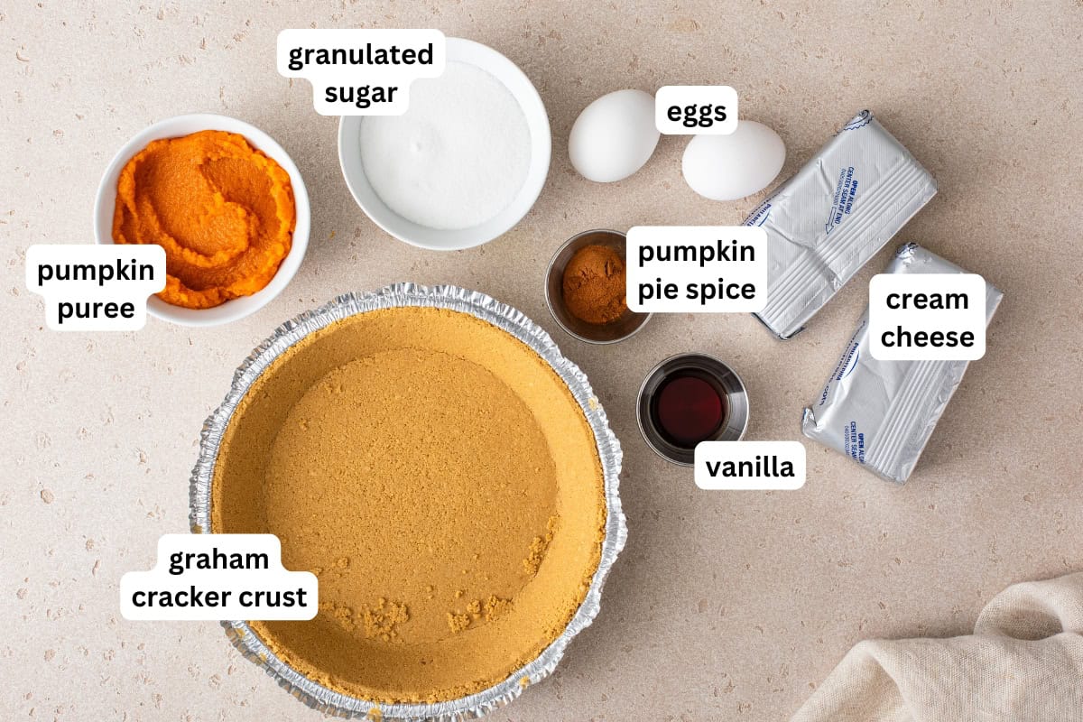 This easy Pumpkin Pie Cheesecake has a hidden cheesecake layer under the spiced pumpkin. Just ONE bowl of batter and no water bath needed! Labeled ingredients for pumpkin pie cheesecake recipe on a countertop: pumpkin puree, granulated sugar, eggs, pumpkin pie spice, cream cheese, vanilla, and a graham cracker crust.