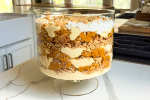 Pumpkin Pie Pudding layers a whole pumpkin pie with buttery graham crackers and the fluffiest pudding into the creamiest no-bake dessert! A layered pumpkin pudding in a trifle dish, including whipped cream, vanilla pudding, crumbled graham cracker pie crust, and pumpkin pie pieces, sits on a marble kitchen countertop with sunlight streaming through the windows in the background.