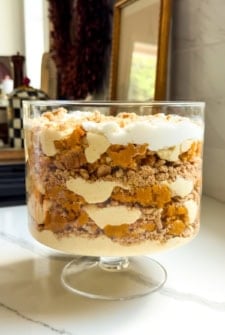 A layered pumpkin pie pudding in a glass trifle bowl, featuring whipped cream, vanilla pudding, crumbled graham cracker crust, and pumpkin pie pieces, sits on a white countertop in a kitchen.
