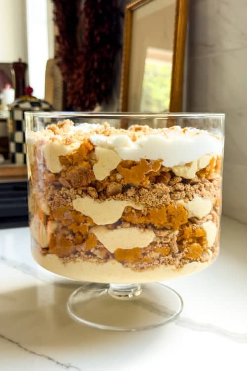 A layered pumpkin pie pudding in a glass trifle bowl, featuring whipped cream, vanilla pudding, crumbled graham cracker crust, and pumpkin pie pieces, sits on a white countertop in a kitchen.