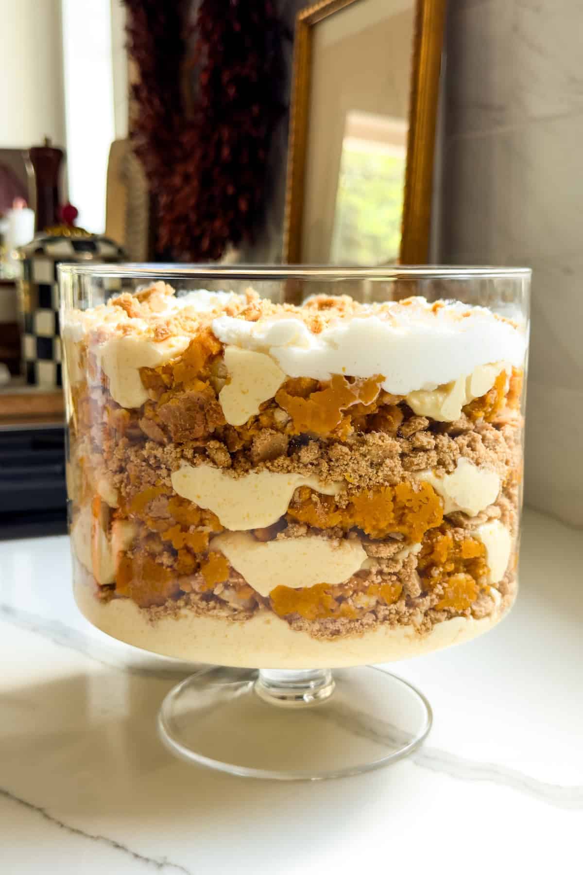 Pumpkin Pie Pudding layers a whole pumpkin pie with buttery graham crackers and the fluffiest pudding into the creamiest no-bake dessert! A layered pumpkin pie pudding in a glass trifle bowl, featuring whipped cream, vanilla pudding, crumbled graham cracker crust, and pumpkin pie pieces, sits on a white countertop in a kitchen.