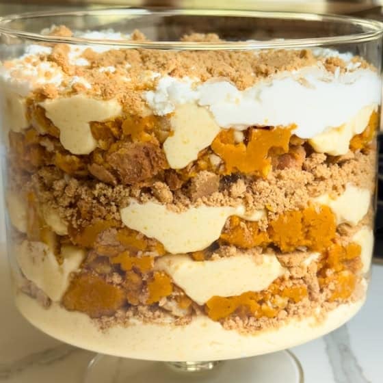 A large glass trifle dish filled with layered pumpkin pie pudding trifle dessert, including whipped cream, vanilla pudding, crumbled graham cracker pie crust, and pieces of pumpkin pie, sits on a marble countertop in a bright kitchen.
