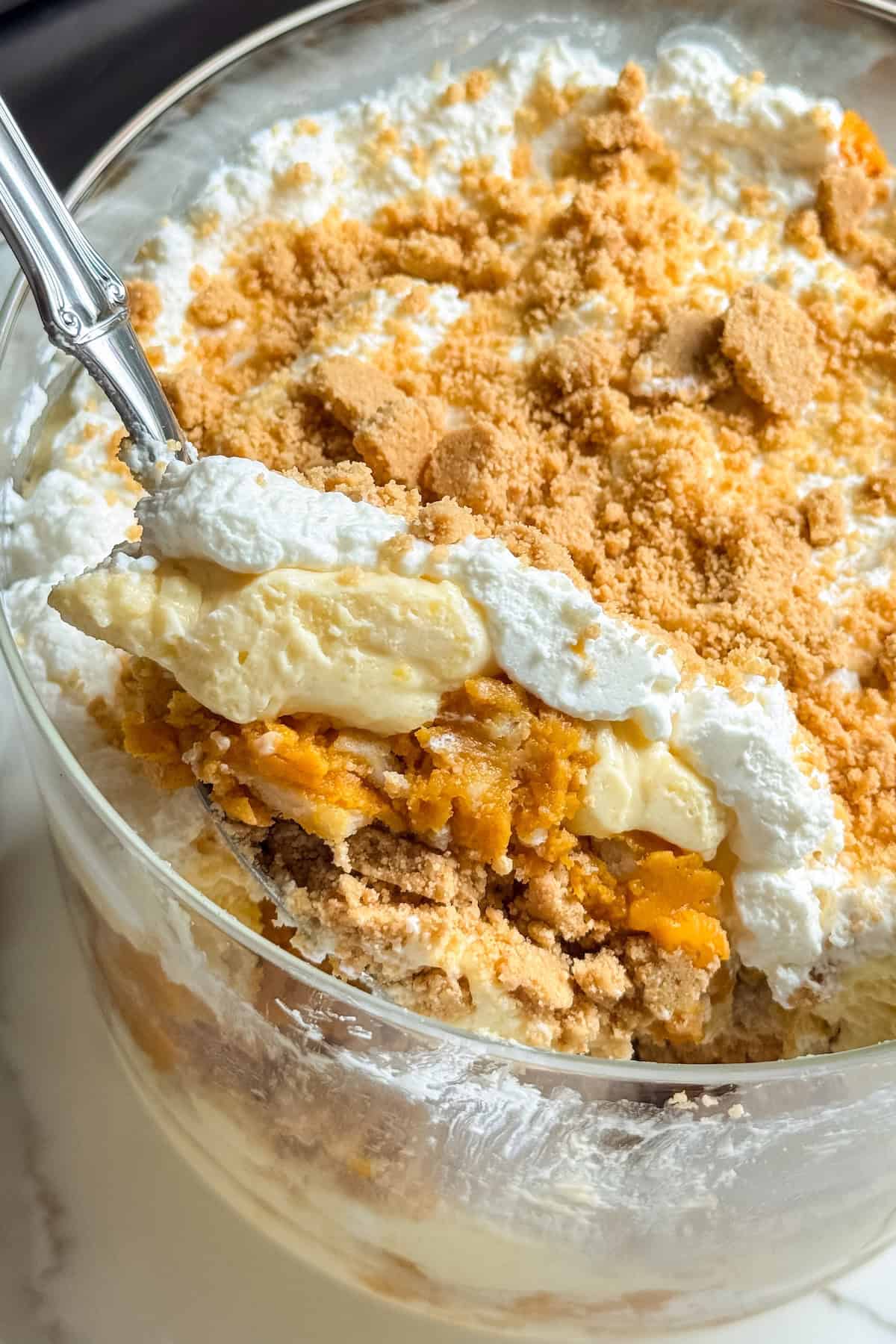 Pumpkin Pie Pudding layers a whole pumpkin pie with buttery graham crackers and the fluffiest pudding into the creamiest no-bake dessert! A close-up of a pumpkin pie pudding trifle with layers of whipped cream, vanilla pudding, crumbled graham cracker crust, and pumpkin pie pieces. A spoon is lifting a serving from the glass dish, showing the pumpkin trifles different textures and layers.