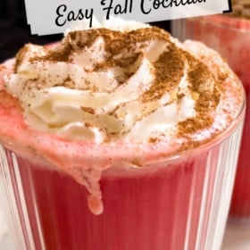 A glass mug of red hot apple cider topped with whipped cream and cinnamon.
