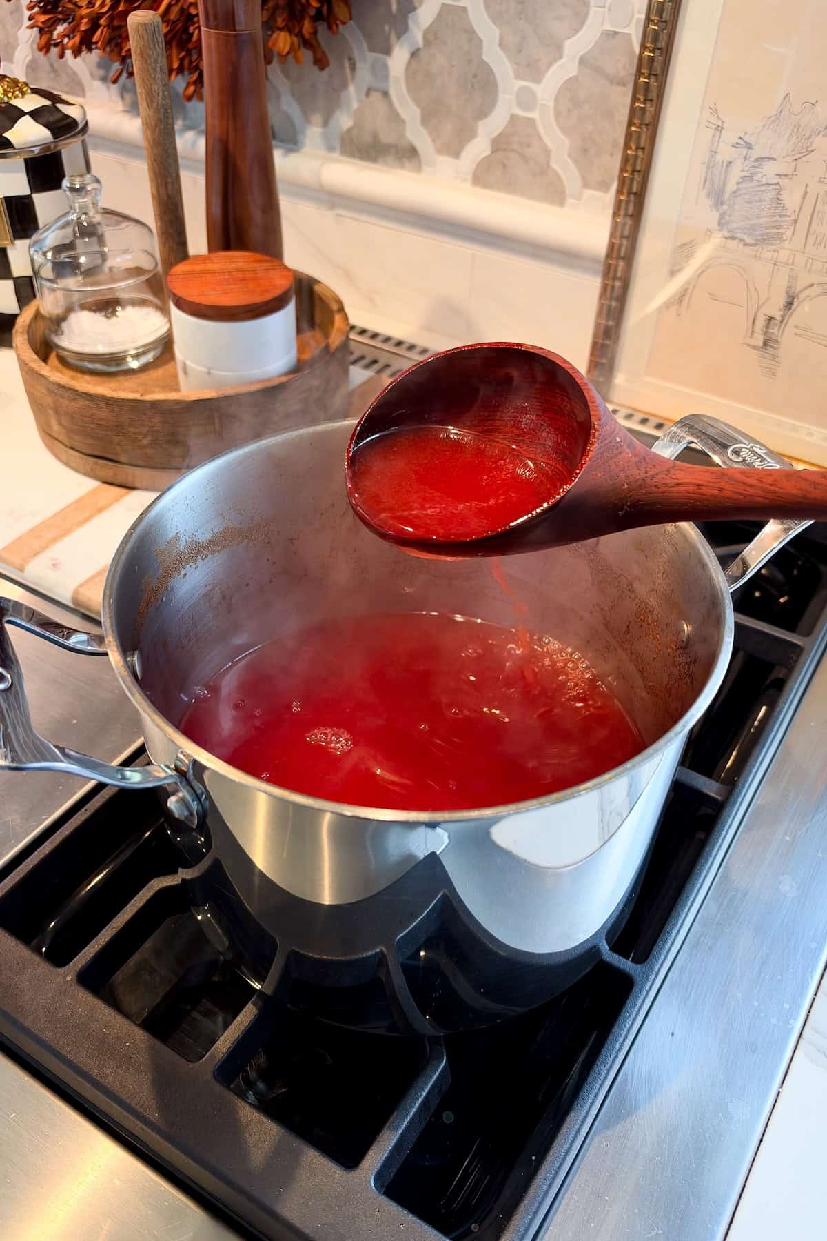 Made with just 2 ingredients, Red Hot Apple Cider tastes like a cinnamon candy apple. Add booze for a dangerously good Apple Cider Cocktail! A wooden ladle lifts vibrant red hot apple cider from a stainless steel pot steaming on a stovetop.