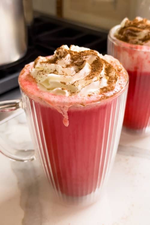 Two fluted glass mugs filled with red hot apple cider, topped with whipped cream and a dusting of cinnamon.