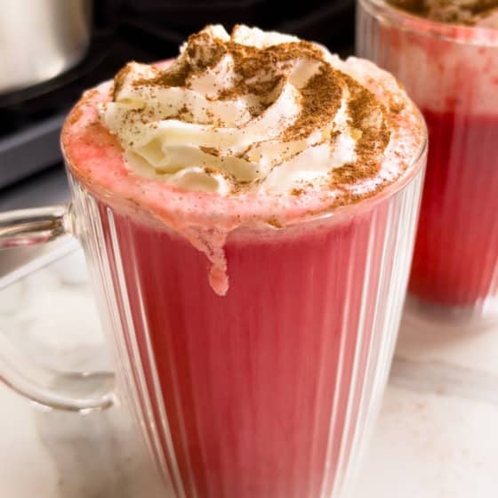 Two fluted glass mugs filled with red hot apple cider, topped with whipped cream and a dusting of cinnamon.