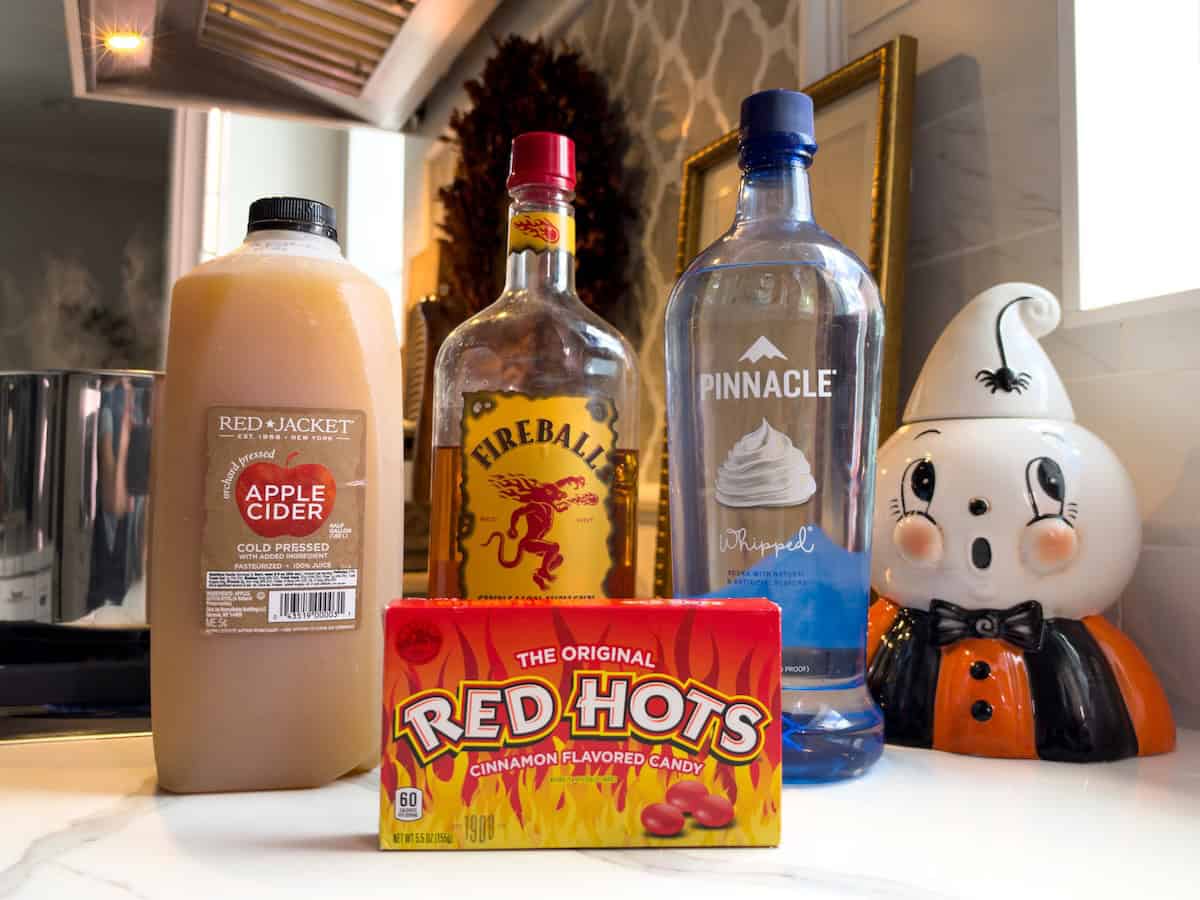 Made with just 2 ingredients, Red Hot Apple Cider tastes like a cinnamon candy apple. Add booze for a dangerously good Apple Cider Cocktail! A jug of Red Jacket apple cider, a bottle of Fireball cinnamon whisky, a bottle of Pinnacle whipped vodka, and a box of Red Hots cinnamon candy in front.