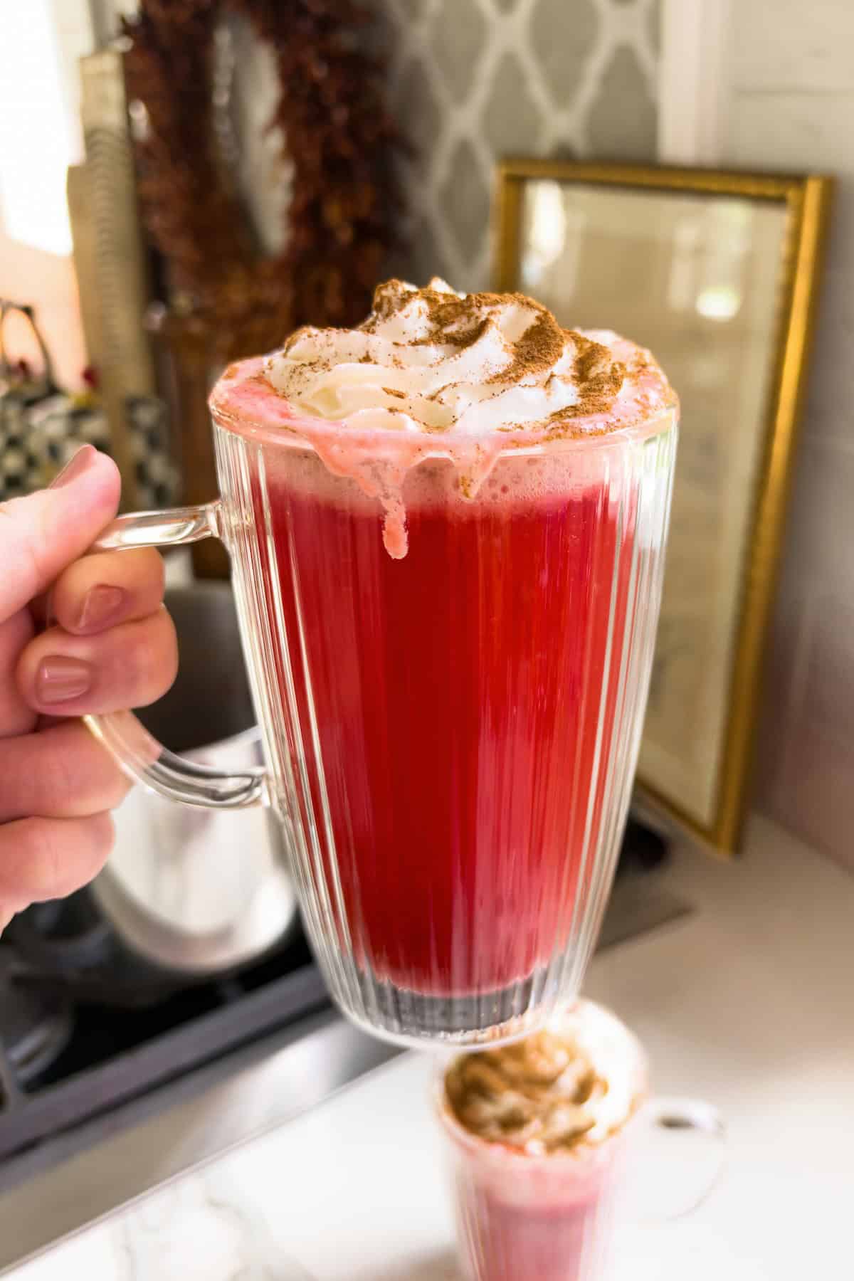 Made with just 2 ingredients, Red Hot Apple Cider tastes like a cinnamon candy apple. Add booze for a dangerously good Apple Cider Cocktail! A hand holds a tall, ribbed glass mug filled with a vibrant red hot apple cider cocktail, topped with whipped cream and a dusting of cinnamon.